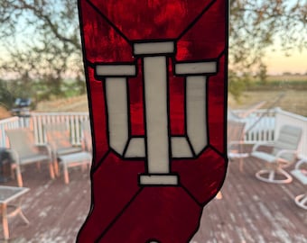 May include: A stained glass piece in the shape of the state of Indiana, with a red background and white "I" symbols. The glass is outlined in black and hangs from a chain. The background shows a deck and outdoor scenery.
