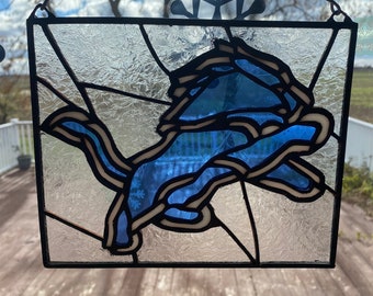 May include: Stained glass window art featuring the Detroit Lions logo. The logo is a blue lion with a white outline and black accents, set against a textured, clear glass background. The window is framed in black and hangs from a chain.