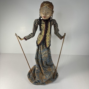May include: A Wayang Golek rod puppet featuring a painted wooden head with a brown headpiece, wearing a striped long-sleeve top with a gold-trimmed black vest and a blue and brown patterned batik sarong, held by two thin wooden control rods.