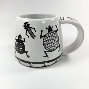 May include: White ceramic mug with black cartoon figures. The mug features three stylized figures with geometric patterns and shapes. The handle is white and the base has a black border. The mug is designed for hot beverages.
