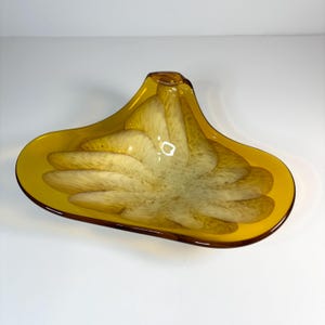 May include: A decorative, amber-colored glass bowl with a unique, organic shape. The bowl features a contrasting interior design of creamy white and amber swirls. The rim is a darker shade of amber, creating a striking visual contrast. This art glass piece is ideal for display.