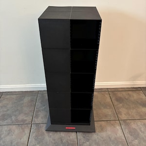 May include: A tall, black storage unit with a trapezoidal shape. The unit is divided into sections, with some appearing to be open shelves. It stands on a rectangular base. The unit is made of a matte black material.