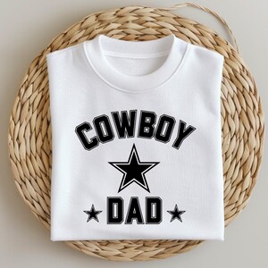 May include: White sweatshirt with black text that reads "COWBOY" in a large font with a black star in the middle and "DAD" in a smaller font with a star on either side.