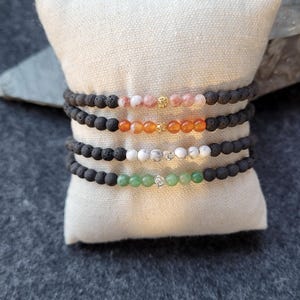 Könnte beinhalten: Vier Armbänder mit Perlen liegen auf einem weißen Kissen. Jedes Armband hat schwarze Lavaperlen und eine Reihe farbiger Edelsteine: rosa, orange, weiß und grün. Goldfarbene Abstandshalter fügen Details hinzu. Die Armbänder werden auf einer dunkelgrauen Oberfläche präsentiert.