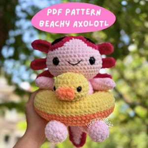 May include: A crocheted axolotl plushie in pink, red, and black, with a yellow and orange inner tube. The axolotl has black eyes and a smiling mouth. The text "PDF PATTERN BEACHY AXOLOTL" is in a pink oval above the plushie.