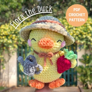 以下が含まれることがあります： 帽子をかぶり、赤いイチゴと灰色のじょうろを持った、かぎ針編みの黄色いアヒル。アヒルはオレンジ色のくちばしと足、ピンクの頬、茶色のリボンをしています。画像には「Flora the Duck」と「PDF Crochet Pattern」と書かれています。
