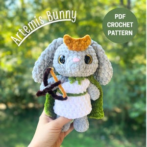 May include: A handmade crochet bunny doll, in shades of gray, white, and green, with a gold crown and a bow and arrow. The doll is wearing a white dress and a green cape. Text on the image reads "Artemis Bunny" and "PDF Crochet Pattern".