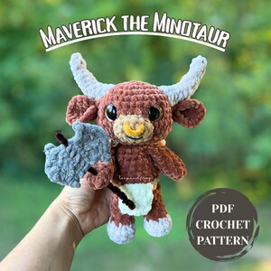 Può includere: Un peluche di minotauro all'uncinetto marrone e bianco fatto a mano con grandi occhi neri, un anello al naso giallo e corna grigie. Il peluche tiene un'ascia grigia e indossa un pannolino bianco. Il testo "Maverick the Minotaur" è sopra il peluche. Il testo "PDF Crochet Pattern" è in un cerchio.