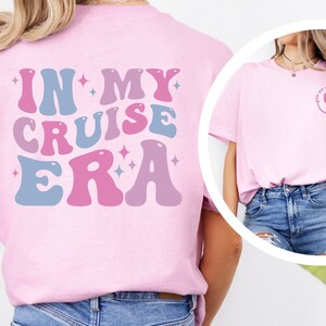 Puede incluir: Camiseta rosa con el texto "In My Cruise Era" en fuente retro azul y rosa con estrellas. La parte posterior de la camiseta tiene una carita sonriente rosa y azul con un rayo.