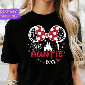 Puede incluir: Camiseta negra con un lazo de lunares rojos y blancos y el texto "Best Auntie ever". El dise&ntilde;o incluye una silueta de castillo y orejas de Mickey Mouse.