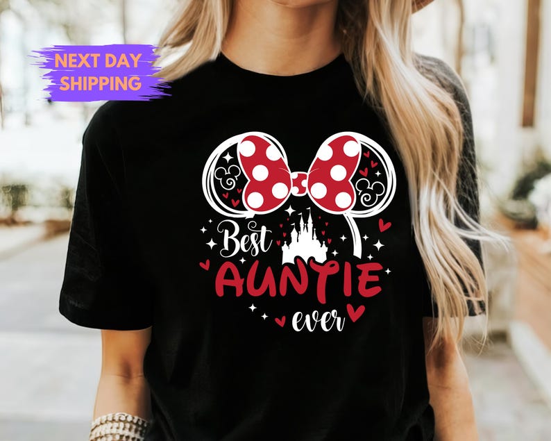 Puede incluir: Camiseta negra con un lazo de lunares rojos y blancos y el texto "Best Auntie ever". El dise&ntilde;o incluye una silueta de castillo y orejas de Mickey Mouse.