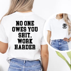 May include: White t-shirt with the text "NO ONE OWES YOU SHIT. WORK HARDER" in bold black letters on the back. A smaller image of a skull with tools is on the front. The shirt is paired with blue jeans.