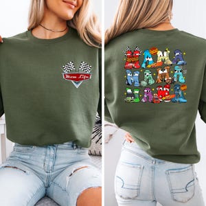 May include: An olive green sweatshirt featuring a "Mom Life" graphic with checkered flags on the front. The back displays a colourful graphic of cartoon cars arranged to form letters.