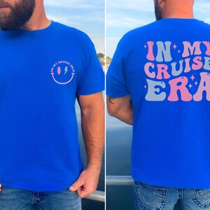 Puede incluir: Una camiseta azul real con una carita sonriente rosa y blanca con un rayo en la parte delantera y el texto "IN MY CRUISE ERA" en rosa y gris en la parte trasera.