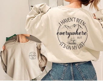 Puede incluir: Una sudadera beige con un estampado gráfico negro que dice "No he estado en todas partes, pero está en mi lista". El gráfico incluye una cordillera y un velero.
