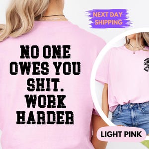 May include: Light pink t-shirt with the text "NO ONE OWES YOU SHIT. WORK HARDER" in bold black letters on the back. The front of the shirt features a small graphic of a skull and wrenches with the text "NOBODY CARES WORK HARDER".