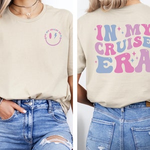 Puede incluir: Una camiseta beige con un gr&aacute;fico de cara sonriente en la parte delantera y el texto "In My Cruise Era" en fuente retro rosa y azul en la parte posterior.
