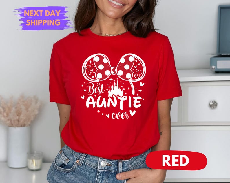 Puede incluir: Camiseta roja con un gr&aacute;fico blanco que presenta un lazo de lunares, un castillo y el texto "Best Auntie ever". La camiseta tiene mangas cortas y cuello redondo. La palabra "RED" se muestra en un &oacute;valo rojo.