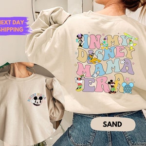 May include: A light beige sweatshirt with a graphic print of Disney characters and the text "In My Disney Mama Era". The sweatshirt is shown on a model wearing blue jeans. The model is standing in front of a white wall.