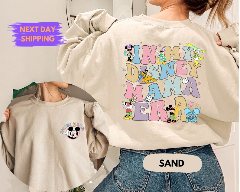 May include: A light beige sweatshirt with a graphic print of Disney characters and the text "In My Disney Mama Era". The sweatshirt is shown on a model wearing blue jeans. The model is standing in front of a white wall.