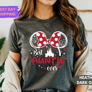 Puede incluir: Camiseta gris oscuro jaspeado con un dise&ntilde;o gr&aacute;fico. El dise&ntilde;o presenta un lazo rojo de lunares, orejas de rat&oacute;n, las palabras "Best Auntie Ever" y una silueta de castillo. La camiseta es de cuello redondo.