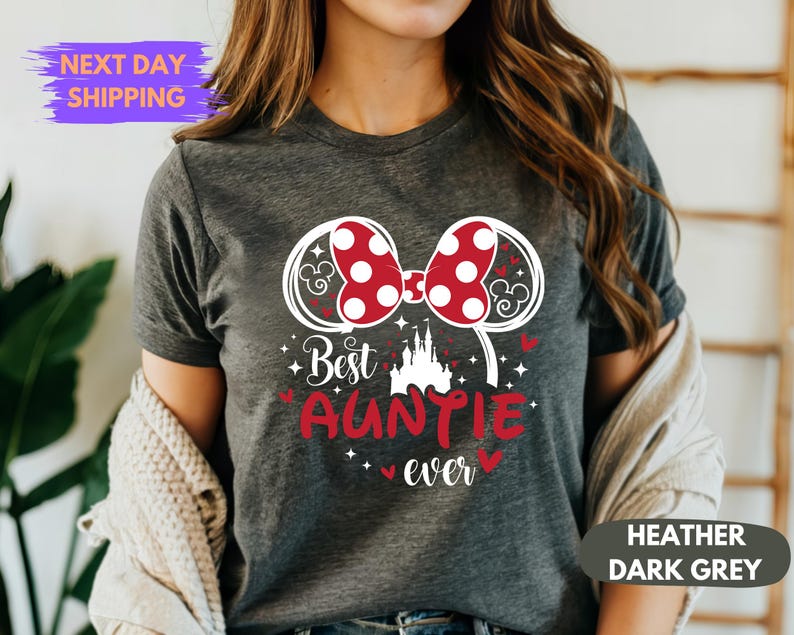 Puede incluir: Camiseta gris oscuro jaspeado con un dise&ntilde;o gr&aacute;fico. El dise&ntilde;o presenta un lazo rojo de lunares, orejas de rat&oacute;n, las palabras "Best Auntie Ever" y una silueta de castillo. La camiseta es de cuello redondo.