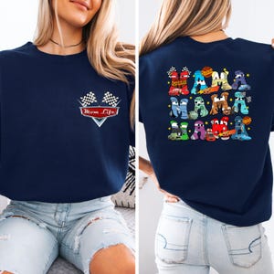 May include: Navy blue sweatshirt featuring a graphic of race cars forming the word "MOM" on the back. The front displays "Mom Life" with checkered flags. A comfortable, casual top.