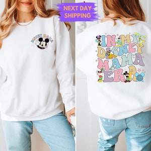 May include: White sweatshirt with a Mickey Mouse graphic on the front and a colorful "In My Disney Mama Era" graphic on the back. The sweatshirt is being modeled by a person wearing blue jeans.
