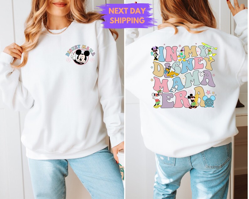 May include: White sweatshirt with a Mickey Mouse graphic on the front and a colorful "In My Disney Mama Era" graphic on the back. The sweatshirt is being modeled by a person wearing blue jeans.