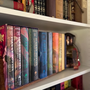 May include: A bookshelf filled with hardcover books, including a series of Harry Potter novels with colorful spines. The books are arranged on white shelves, with other books and decorative items visible.