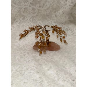 Pode incluir: Uma escultura de árvore de arame com inúmeros pequenos fragmentos de pedra translúcidos de cor âmbar claro fixados em ramos de arame dourado fino e torcido, ancorada numa base de pedra de quartzo rosa pálido e irregular.
