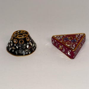 May include: Two decorative trinket boxes. One is a black, cone-shaped box with gold trim and mirrored accents. The other is a triangular, red box with gold trim, mirrored accents, and blue and gold circular designs.