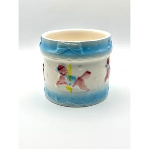 May include: A ceramic container with a light blue bow and ribbon design around the top and bottom. The white center features carousel horse figures in pink, blue, and yellow. The interior is a cream color.