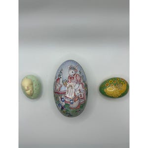 May include: Three decorative Easter egg ornaments. One is light green with a face, the second is oval-shaped with a bunny family scene, and the third is green with yellow flowers and a bunny.