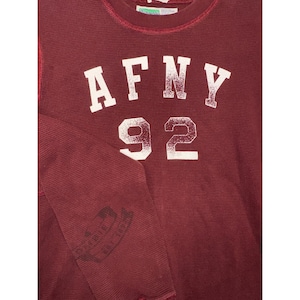May include: Maroon waffle-knit long-sleeved top with the text "AFNY 92" in white block letters. The top has a crew neck and a small graphic on the left sleeve. The fabric has a textured appearance.