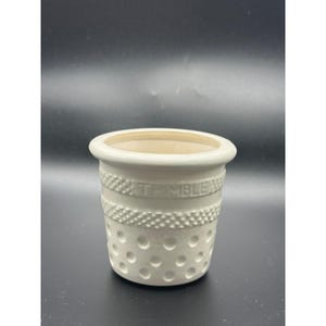 May include: A white ceramic cup shaped like a sewing thimble. The exterior features a textured band with the embossed word THIMBLE, a cross-hatched pattern, and a series of circular indentations around the base, mimicking a classic thimble design.