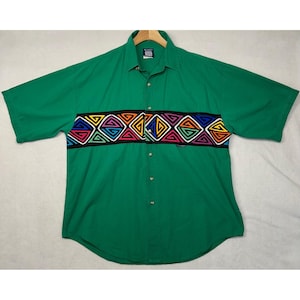 May include: Green short-sleeved shirt with a button-down front. A vibrant geometric pattern in black, yellow, blue, red, and pink adorns a band across the chest. The shirt has a collar and a small pocket.