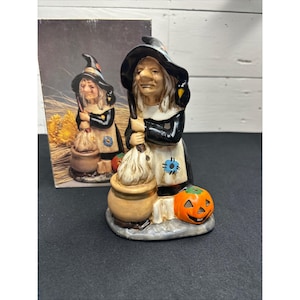 May include: A ceramic witch figurine wearing a black hat and robe with a white apron, stirring a tan cauldron with a broom. A black bird sits on the witch's shoulder, and an orange jack-o'-lantern with a carved face rests at the base of the figure.
