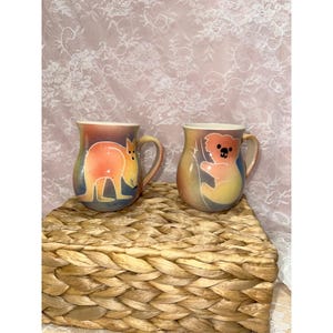 May include: Two ceramic mugs with animal designs. One mug features a kangaroo, the other a koala. Both mugs have a colourful, gradient background with orange, yellow, and blue hues. The mugs are placed on a woven basket.