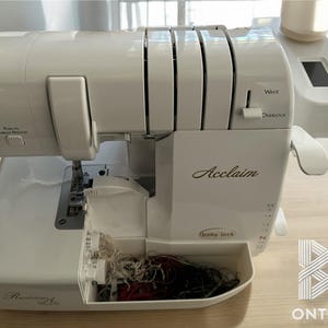 Può includere: Macchina da cucire overlock Acclaim bianca con un vassoio per il filo in plastica trasparente pieno di bobine di filo. La macchina ha un display digitale e una leva per selezionare tra punti a onda e a punto overlock.