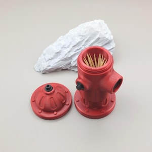 May include: A red, 3D-printed toothpick holder shaped like a fire hydrant, with a matching lid. The holder is open, revealing toothpicks. A white, textured decorative rock is in the background.