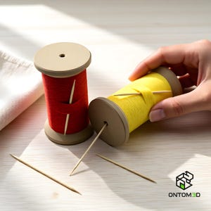 Puede incluir: Dos carretes de hilo, uno rojo y otro amarillo, con palillos de dientes de madera insertados. Los carretes tienen una base y una parte superior beige. Un carrete está en posición vertical, mientras que el otro lo sostiene una mano. El fondo es una superficie de madera clara.
