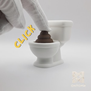 Może przedstawiać: Biała ceramiczna toaleta z brązową emotikoną kupy. Słowo "CLICK" jest napisane na żółto. Obraz jest na białym tle.