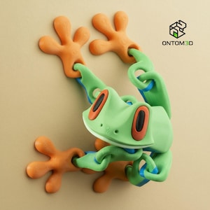 Könnte beinhalten: Eine 3D-gedruckte grüne Froschfigur mit orangefarbenen Füßen und rot umrandeten Augen. Der Frosch ist durch grüne und blaue Glieder verbunden. Das Logo "ONTOM3D" befindet sich in der oberen rechten Ecke. Ein verspieltes Dekorationsobjekt.