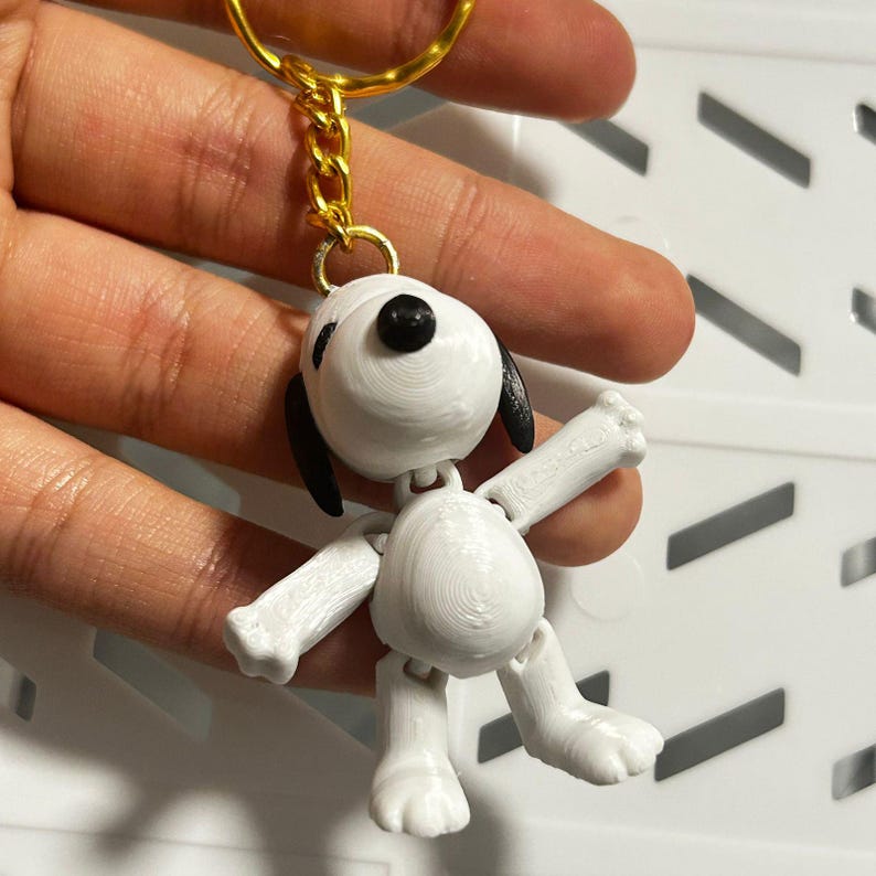 Op de afbeelding: Een witte 3D-geprinte sleutelhanger van Snoopy, de iconische beagle uit de Peanuts-strip. De sleutelhanger heeft Snoopys kenmerkende slappe oren, zwarte neus en zwarte ogen. De sleutelhanger is bevestigd aan een gouden ketting en ring.