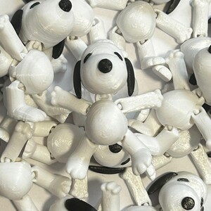 Op de afbeelding: Een groep witte, scharnierende plastic beeldjes van het cartoonpersonage Snoopy. De beeldjes zijn in verschillende poses geplaatst met hun armen en benen gebogen bij de gewrichten.