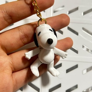 Op de afbeelding: Witte plastic sleutelhanger met een cartoon hond met zwarte ogen en neus. De hond staat met zijn armen en benen uitgestrekt.