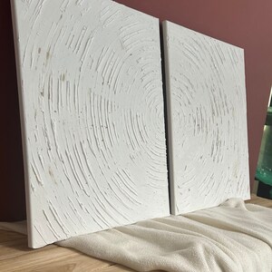 May include: Two white canvas paintings with a textured, circular pattern. The paintings are leaning against a wooden surface.