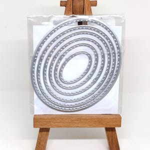 May include: A set of silver-colored metal oval cutting dies nested together. Each die features a decorative stitched edge pattern. The dies are contained within a clear plastic sleeve and are displayed on a small wooden easel stand.