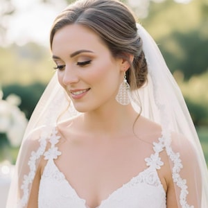 Puede incluir: Una novia con un vestido de novia de encaje blanco y velo. Lleva pendientes colgantes con un diseño calado delicado. Su cabello está peinado en un moño y tiene maquillaje natural. El fondo está borroso, con vegetación y luz solar suave.